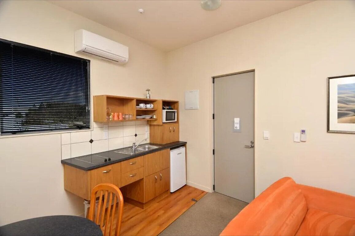 A compact kitchen with a microwave, sink, and a small refrigerator. There is a dining table with chairs and a single orange couch.