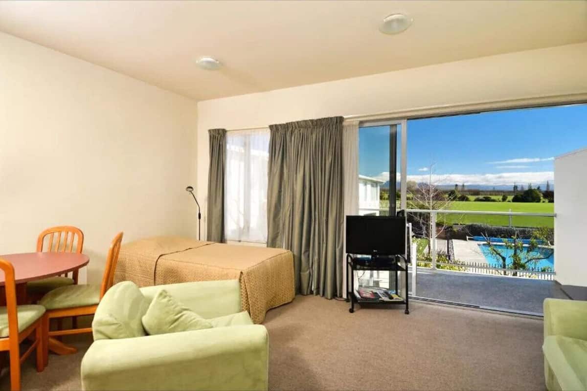 Habitación de hotel con cama, una mesa de comedor con sillas, televisión y balcón con vista a una piscina y montañas.