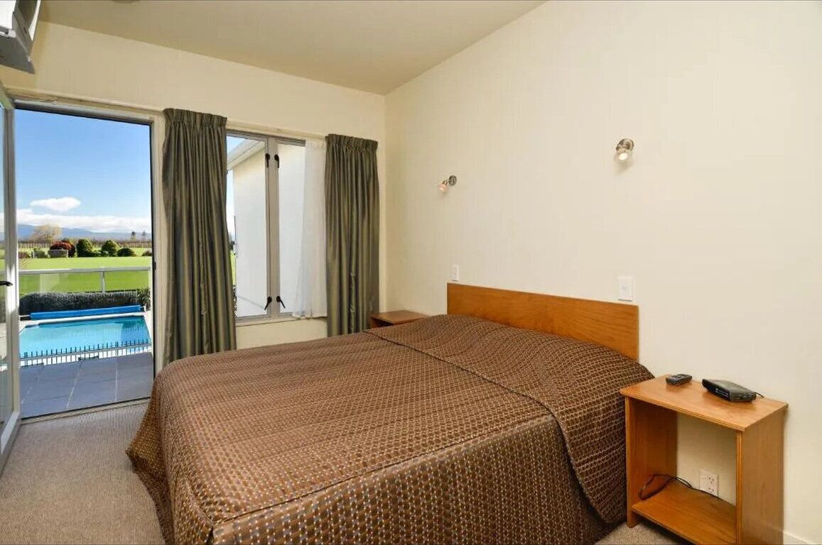 Habitación de hotel con una cama grande, una mesita de noche de madera y vista a una piscina a través de un ventanal.