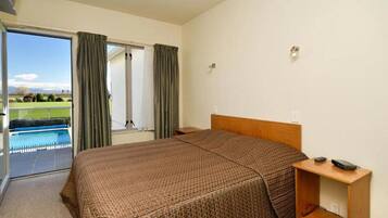 Quarto de hotel com uma cama grande, mesa de cabeceira de madeira e vista para uma piscina através de uma porta de vidro.