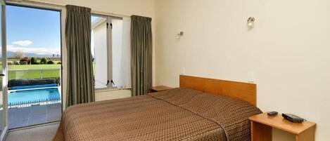Habitación de hotel con una cama grande, una mesita de noche de madera y vista a una piscina a través de un ventanal.