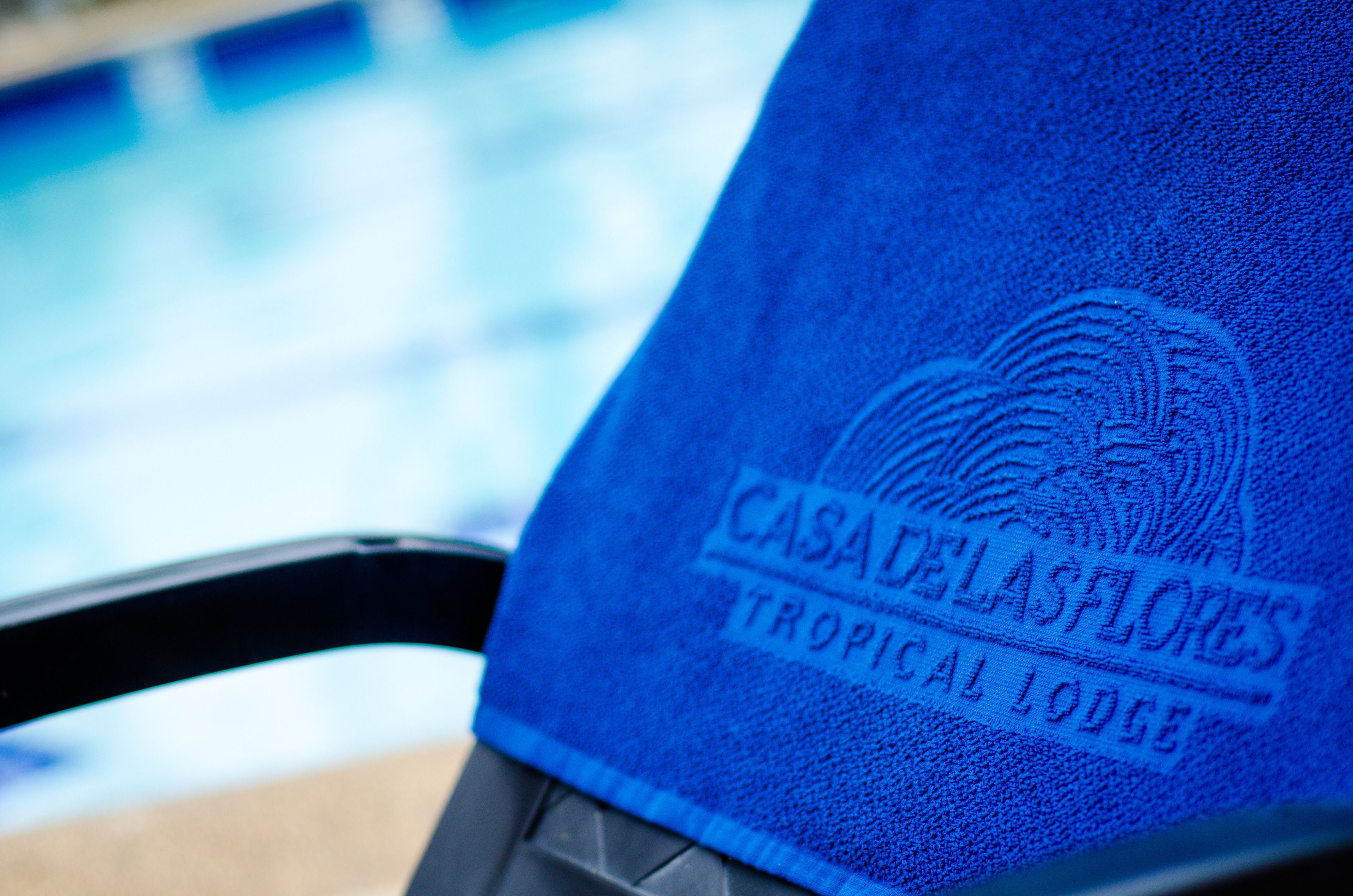 A blue towel with the text "CASA DELAS FLORES TROPICAL LODGE" on a poolside chair.
