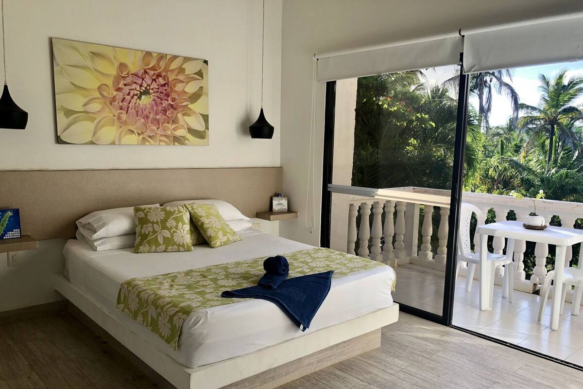 Une chambre à coucher comprenant un lit, un tableau, une table de chevet, une commode, un balcon avec une table et des chaises, et une vue sur la verdure.