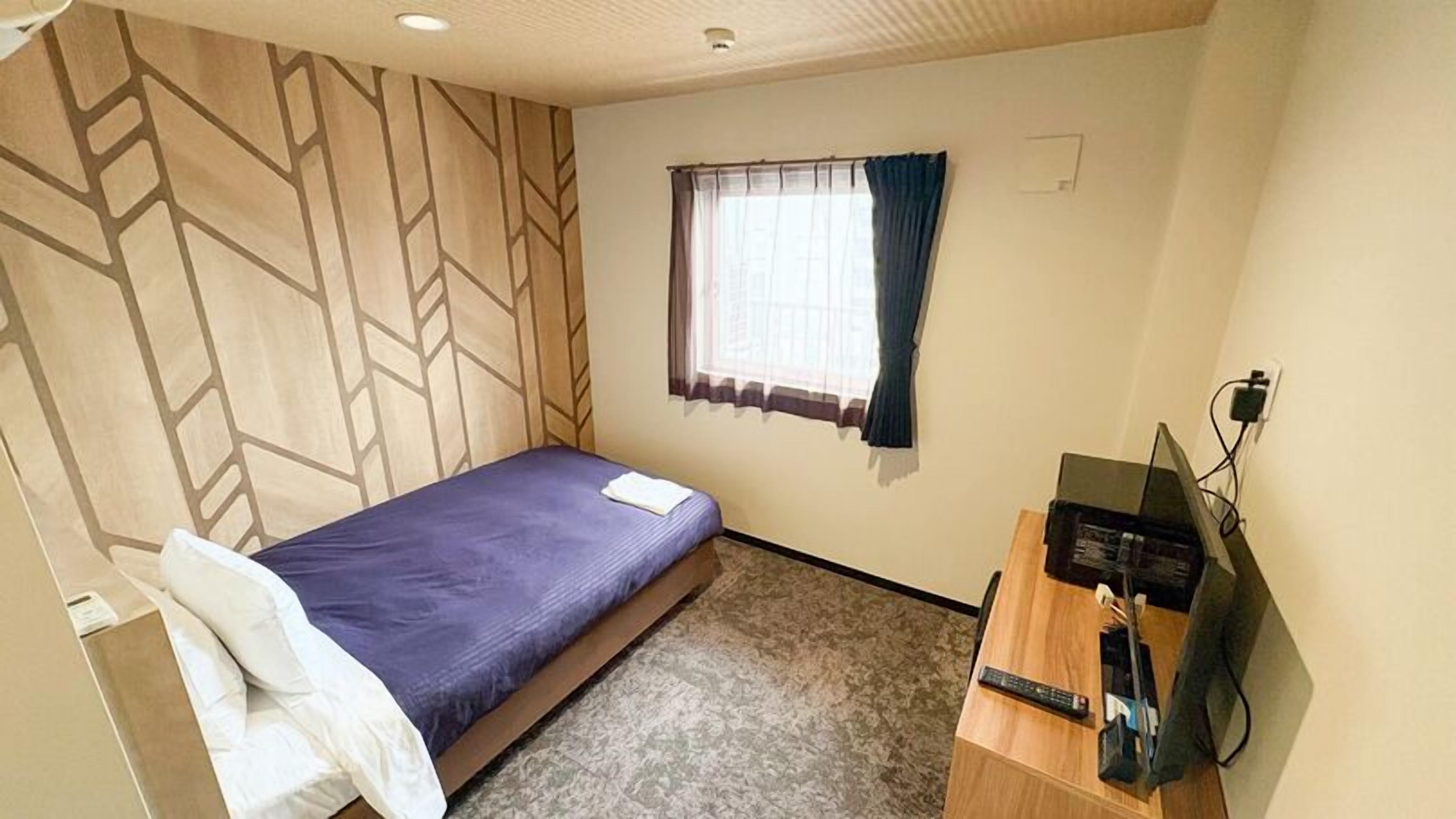 Une petite chambre avec un lit simple, une tête de lit en bois, une fenêtre avec des rideaux, un bureau avec un ordinateur et un téléviseur.