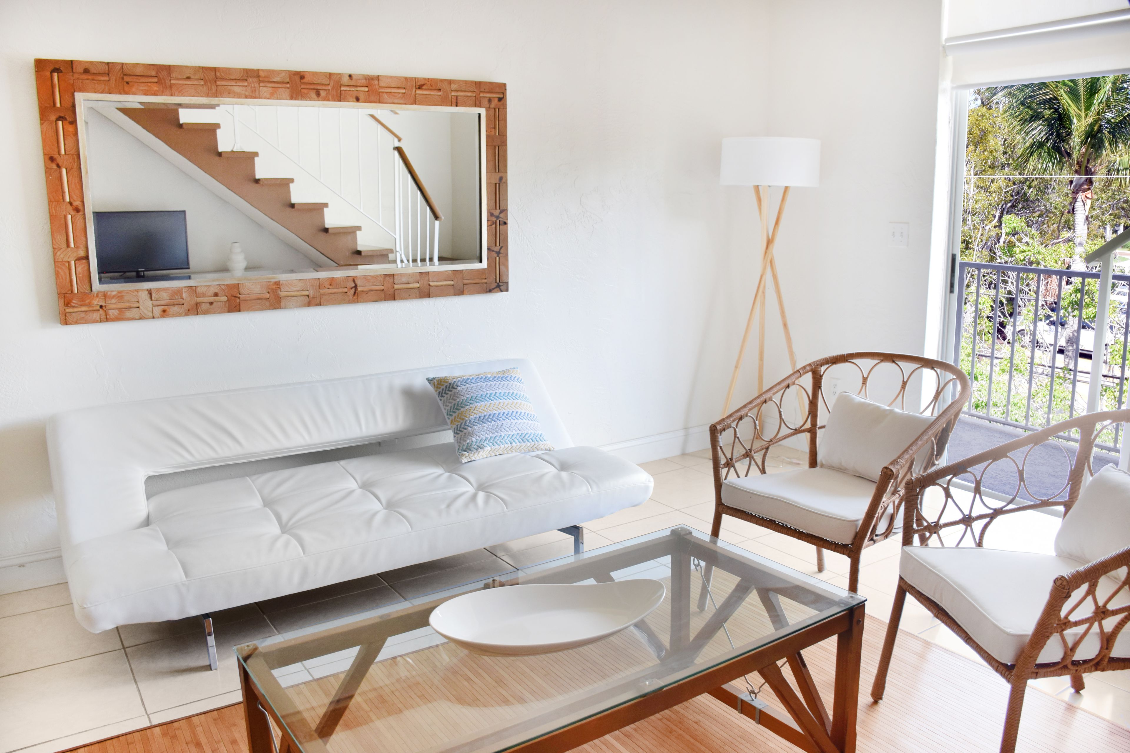 A modern living room with a white sofa, a glass coffee table, wicker chairs, a large mirror, and a view of a staircase.