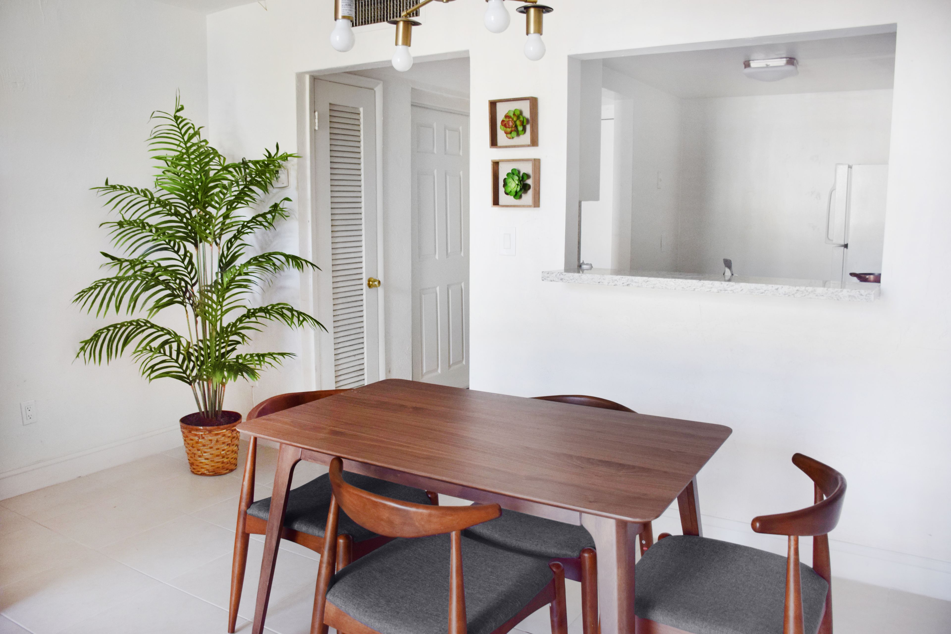 A dining area with a wooden table and chairs, a potted plant, and a white wall with framed pictures.