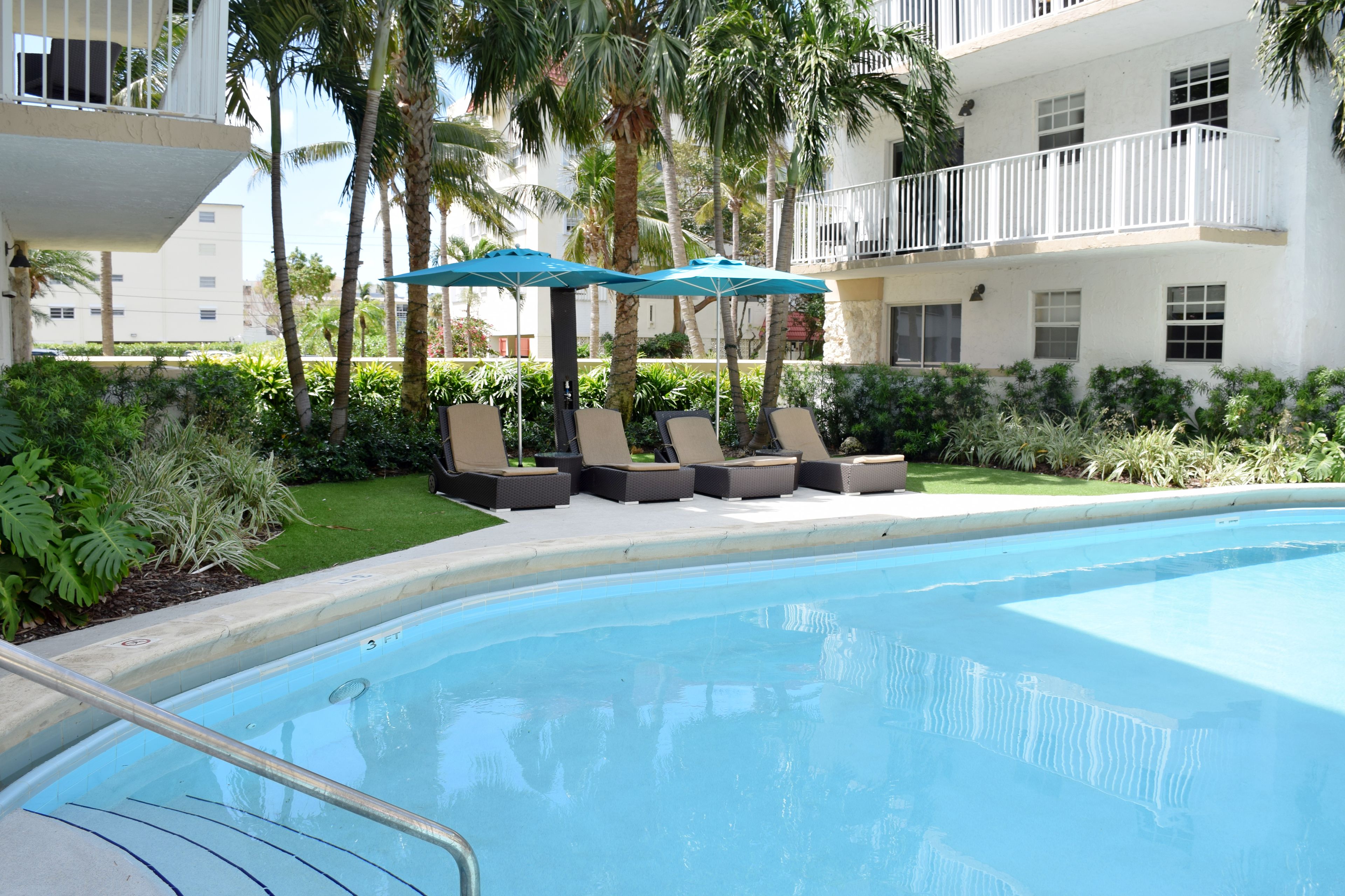 A swimming pool with lounge chairs and umbrellas, surrounded by palm trees and a white building.