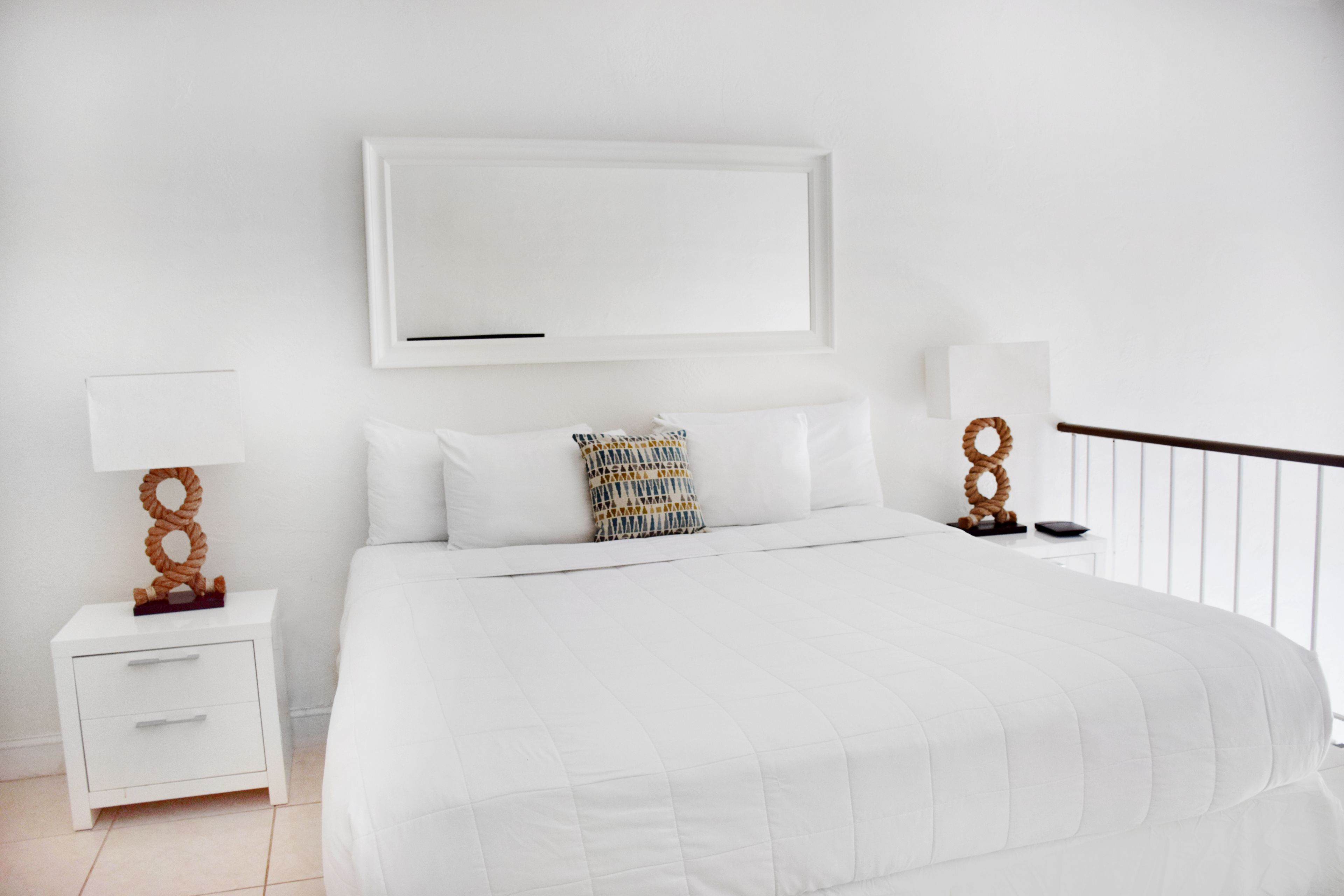 A white bedroom with a bed, two bedside tables, a mirror, and a window with a view.
