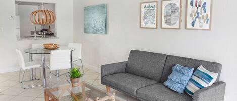 A modern living room with a grey sofa, a glass coffee table, and a dining area with a glass table and chairs.