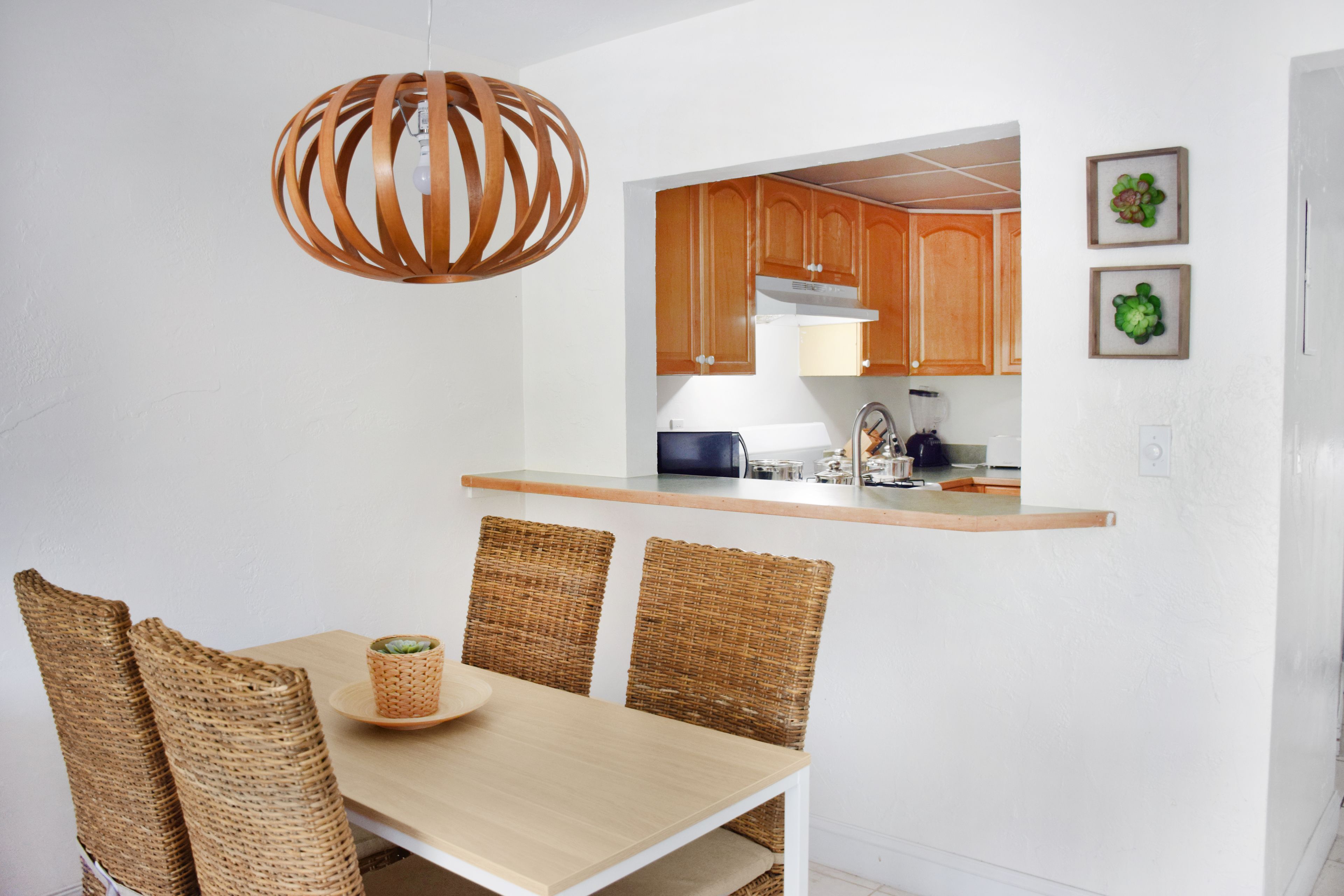 A dining area with a wooden table and wicker chairs, a kitchen with wooden cabinets, and a hanging light fixture.
