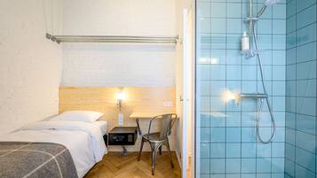 A hotel room with a bed, a desk with a chair, a small table, a nightstand, a wall-mounted heater, and a shower area with blue tiles.