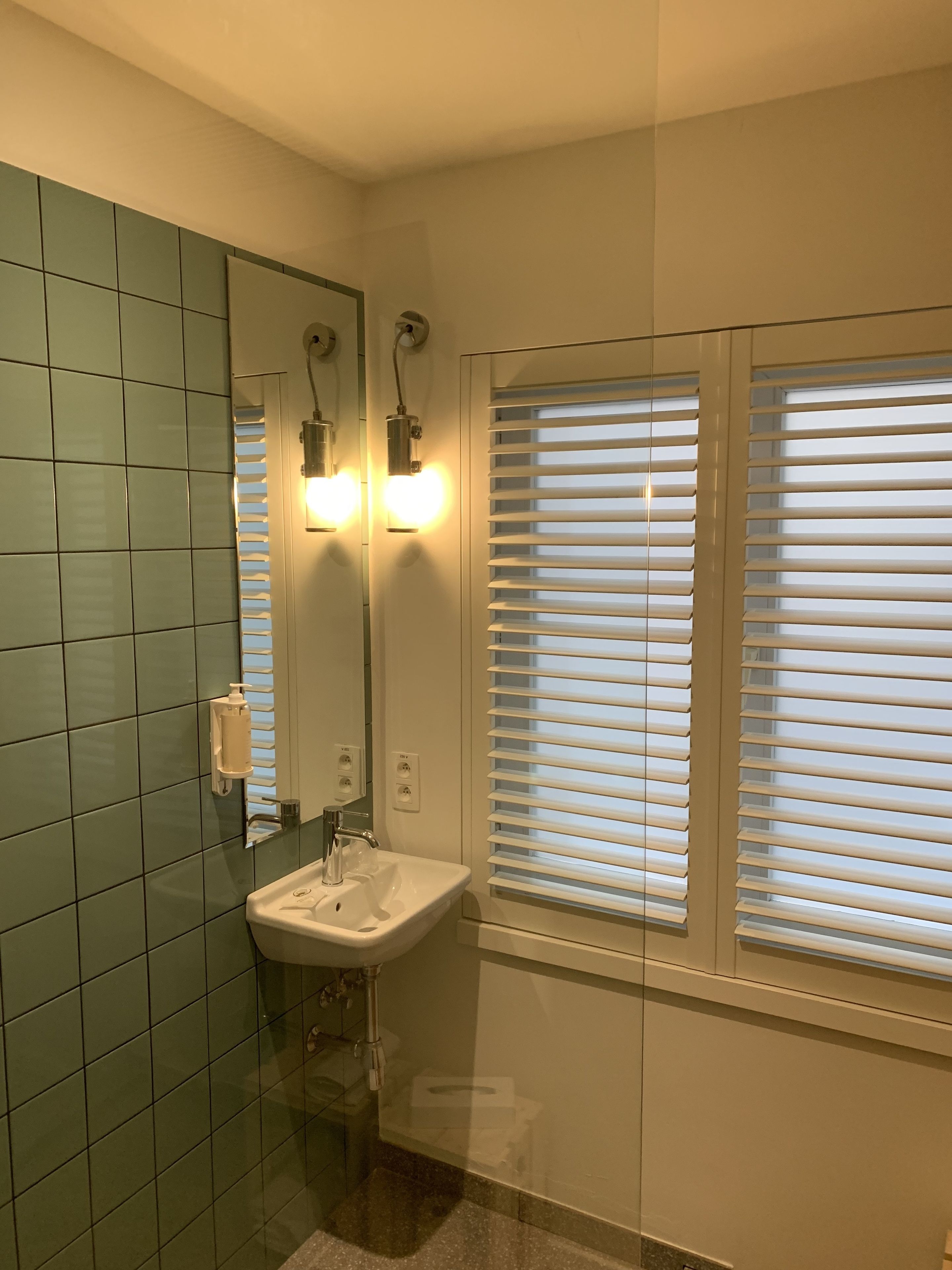 Une salle de bain avec des carreaux verts, un lavabo, un miroir et deux fenêtres équipées de stores.