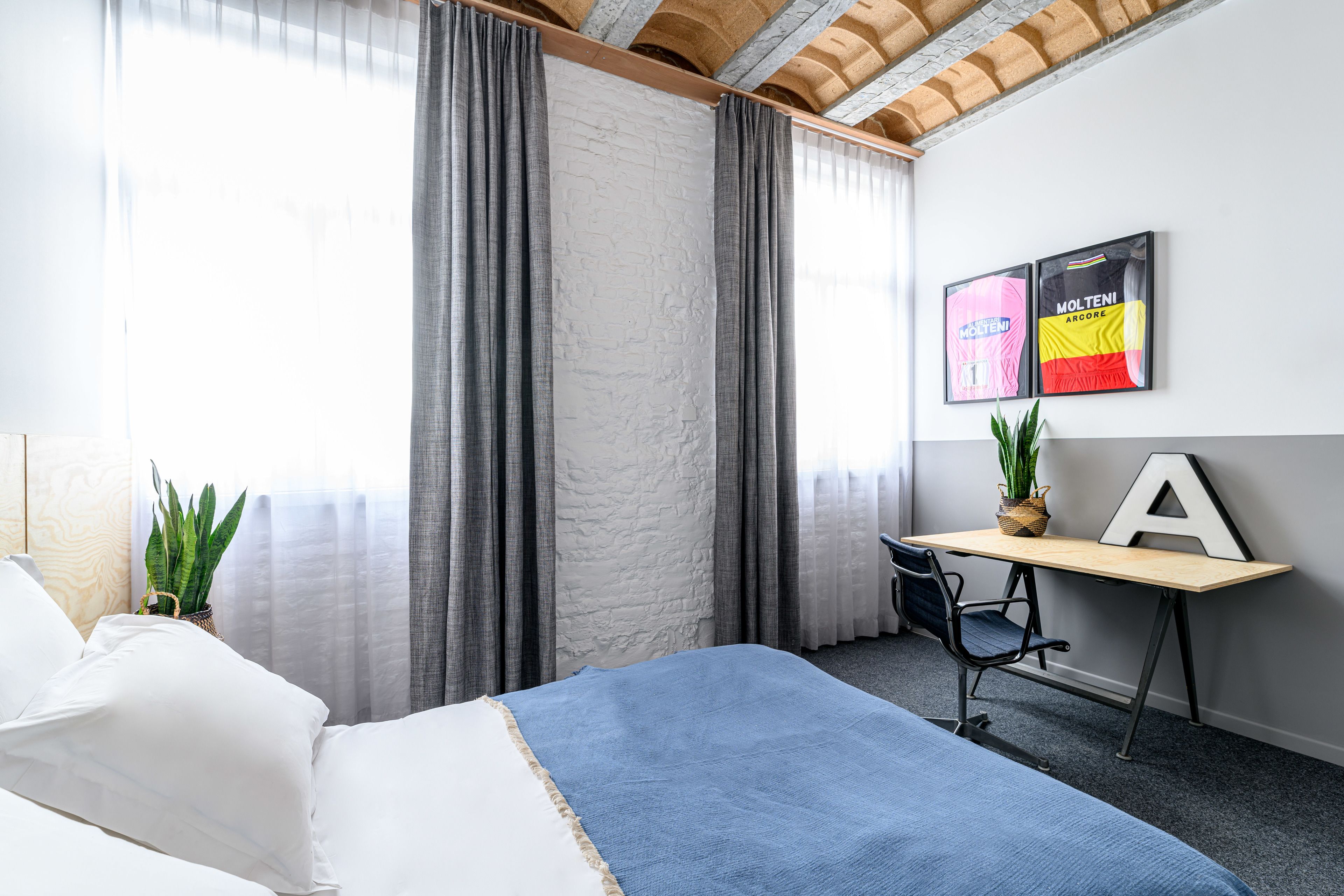 Hotelkamer met een bed, een bureau met stoel, een potplant en twee ingelijste shirts aan de muur.