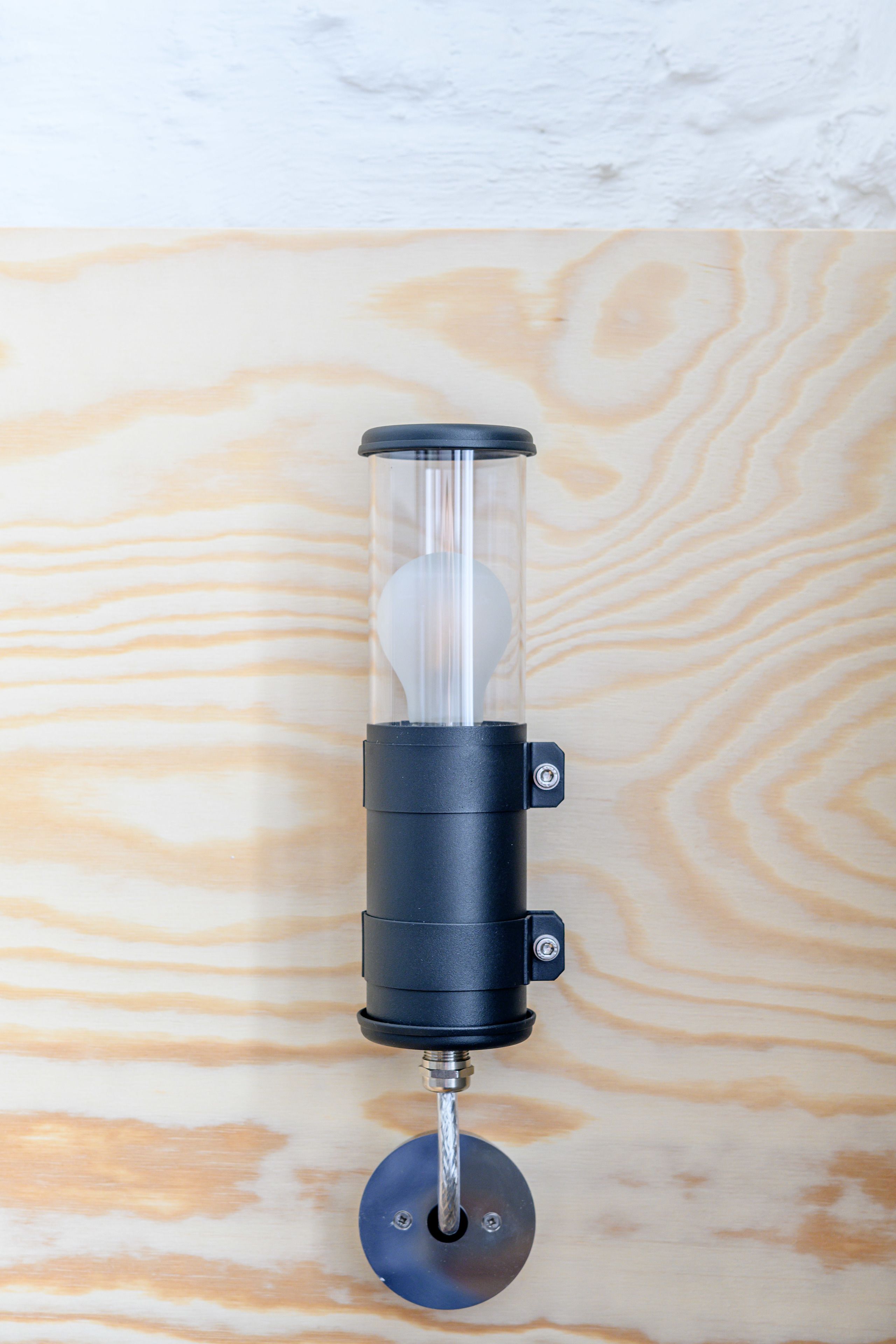 Un luminaire mural fixé au mur, doté d’un abat-jour en verre transparent et d’un socle noir, installé sur un mur en bois présentant un motif ondulé.