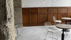 A room with a white brick wall, wooden paneling, and a speckled floor. There are two metal wire chairs and a round table with a black base. - Hotel La Grande Cloche (Brussels)