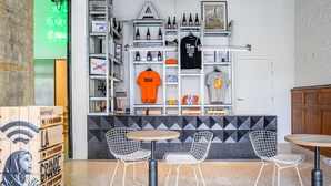 A café with a counter, shelves with bottles, a t-shirt hanging, and two chairs. - Hotel La Grande Cloche (Brussels)