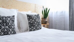A neatly made bed with white bedding and geometric patterned pillows, a wooden headboard, a small side table with a plant, and sheer curtains. - Hotel La Grande Cloche (Brussels)