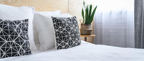 A neatly made bed with white bedding and geometric patterned pillows, a wooden headboard, a small side table with a plant, and sheer curtains.