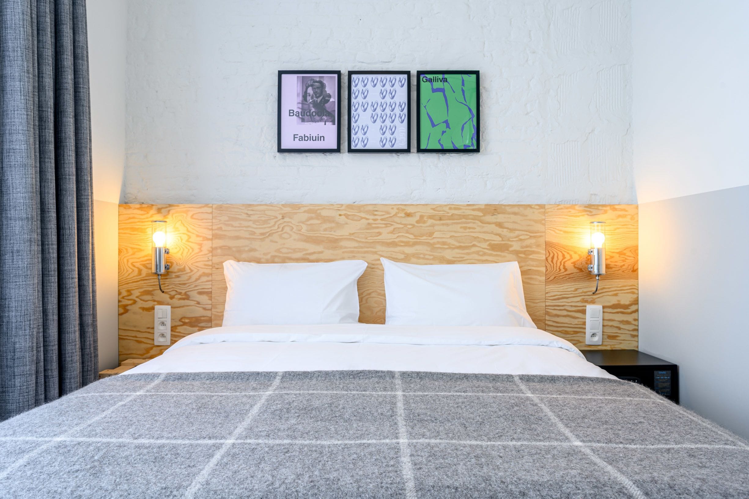 Een net opgemaakt bed met een geruit plaid, twee wandlampen en drie ingelijste foto’s aan de muur.