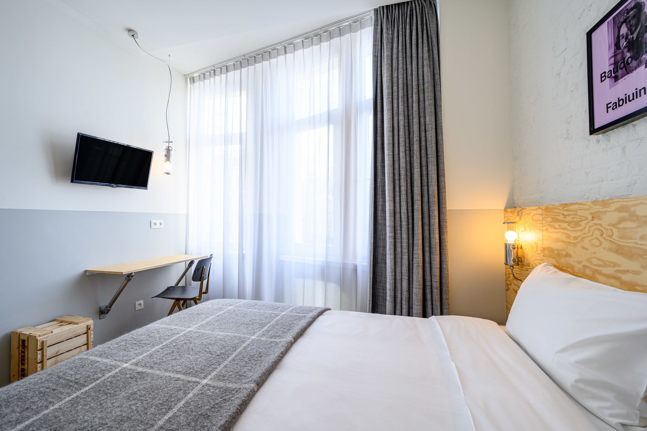 Habitación de hotel con una cama, un escritorio con silla, un televisor montado en la pared y un ventanal con cortinas.