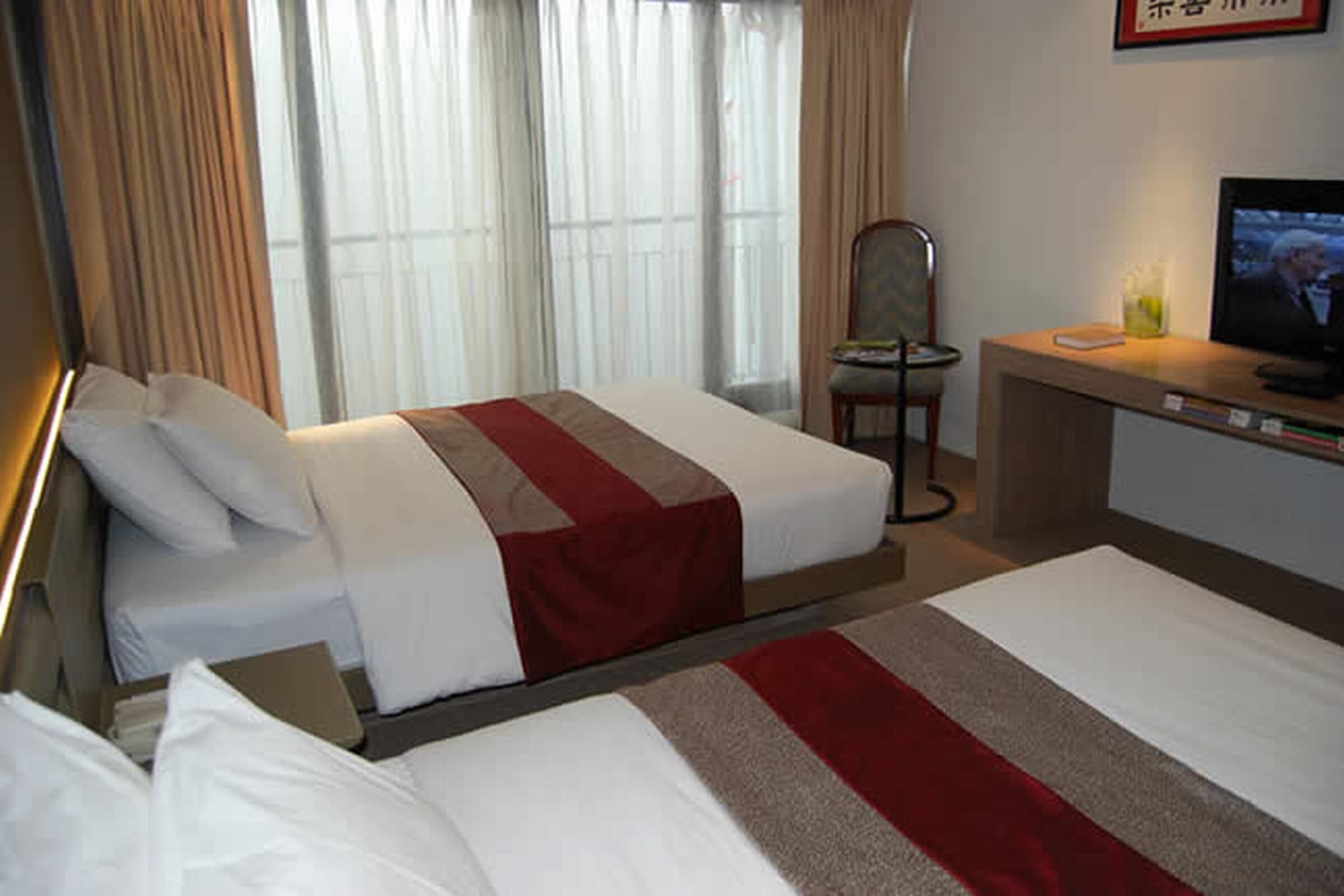 A hotel room with two beds, a desk with a television, a chair, and a window with curtains.