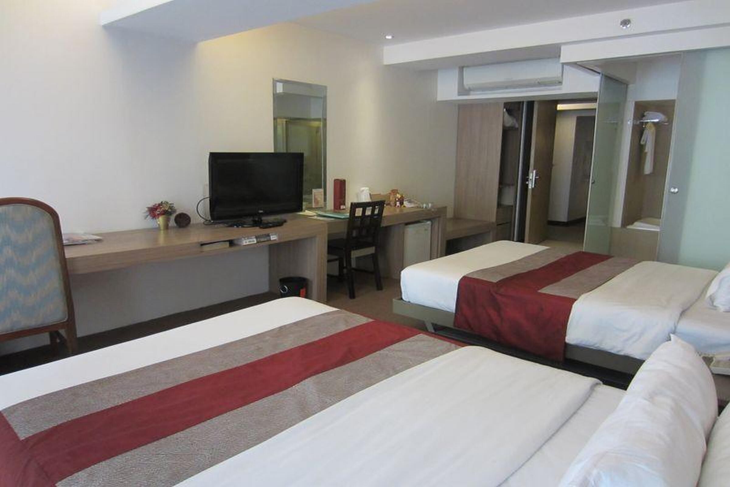 A hotel room with two beds, a desk with a television, a chair, and a shower area.