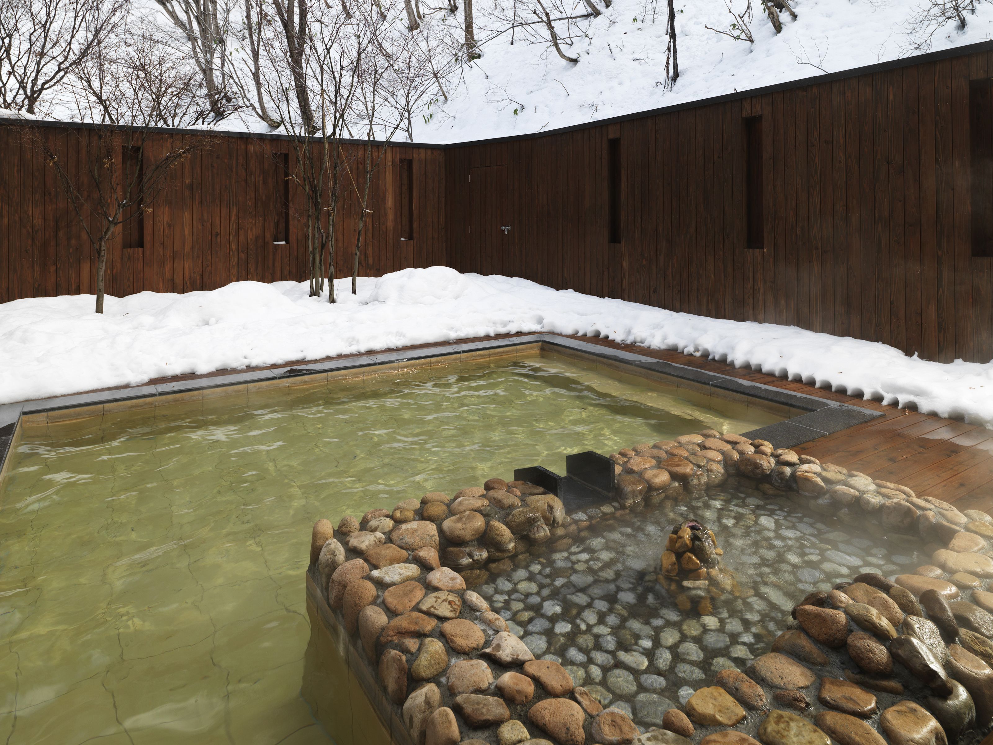 Una piscina rectangular con borde rocoso, rodeada por una terraza de madera y suelo cubierto de nieve.