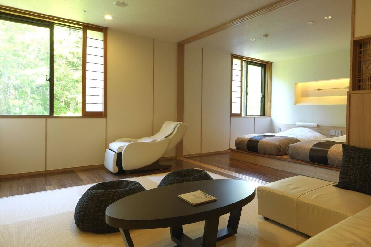 Ein modernes Hotelzimmer mit einer Couch, einem Massagesessel, einem Bett mit Kissen, einem kleinen Tisch und großen Fenstern.