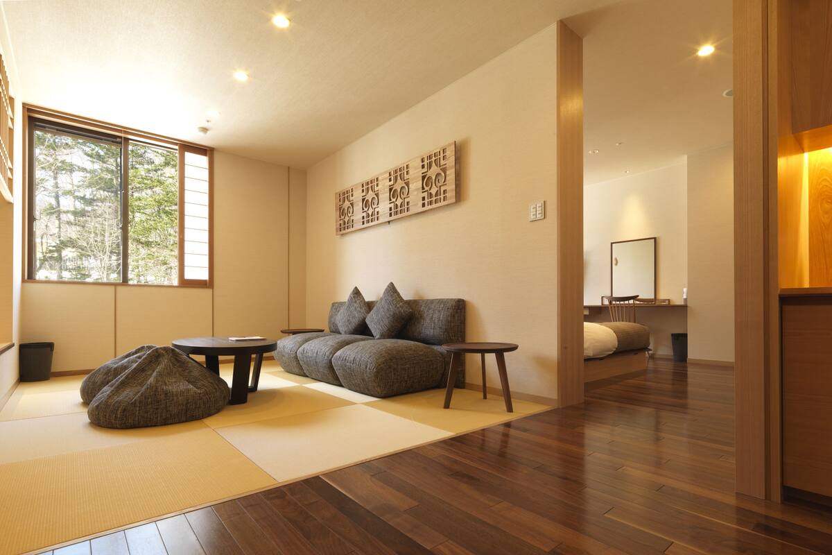 Ein modernes Wohnzimmer mit einer Couch, zwei kleinen Tischen und einem Sitzsack. Ein großes Fenster bietet Blick auf Bäume.