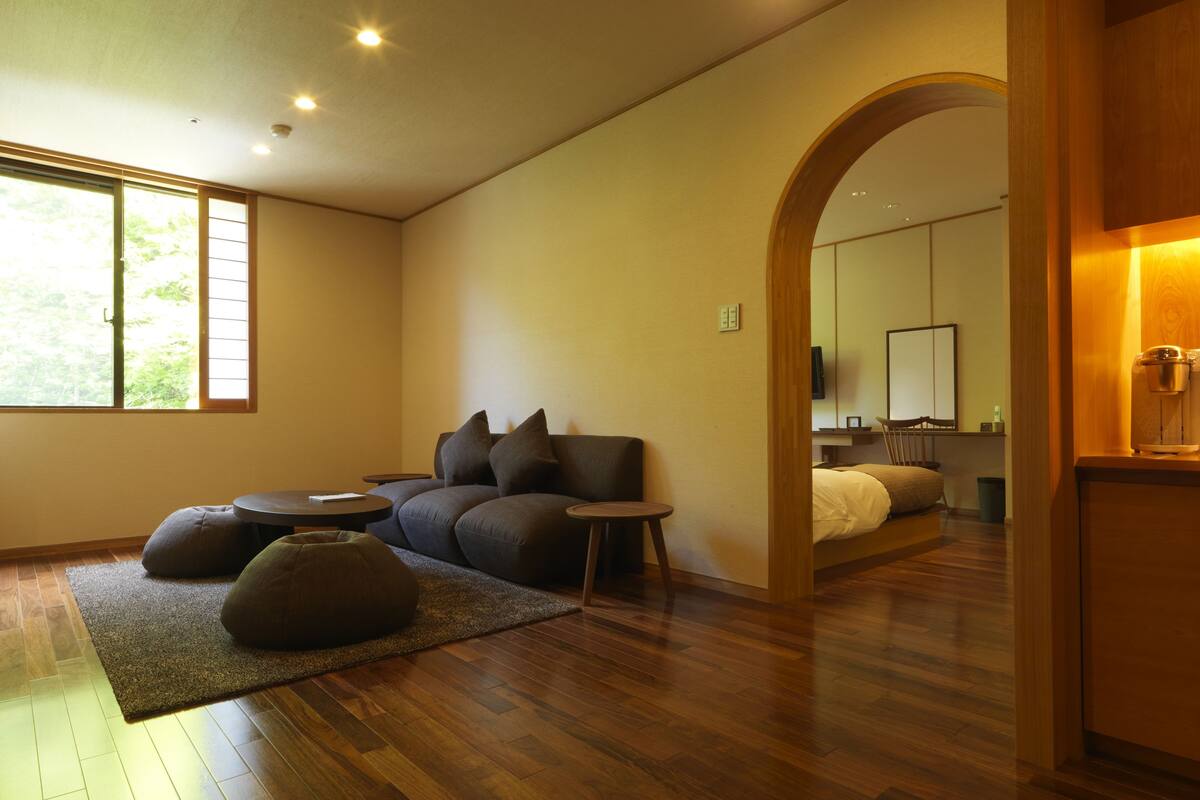 Ein modernes Wohnzimmer mit Sofa, Hocker und Couchtisch, Holzboden und einem großen Fenster.