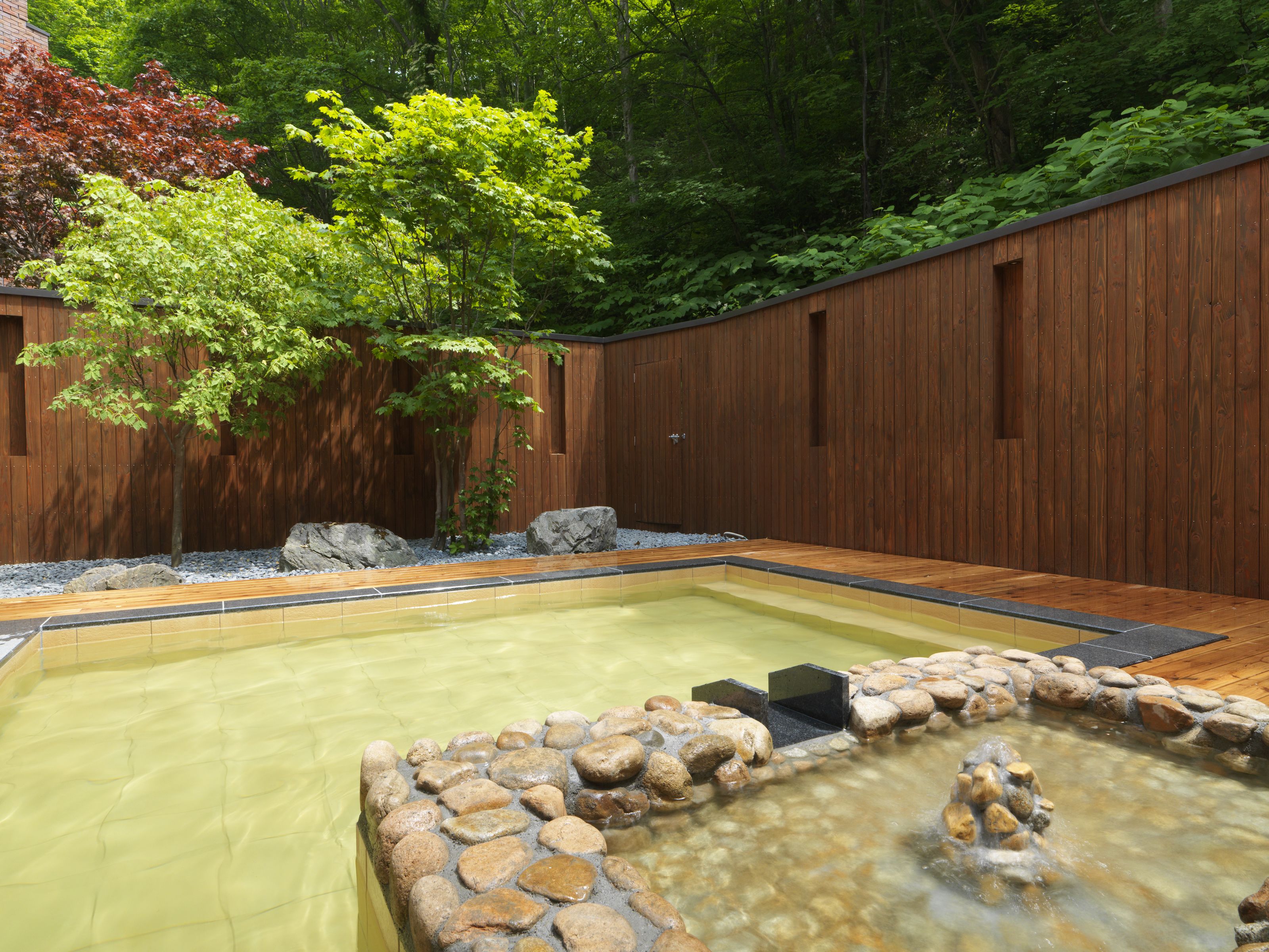 A rectangular hot tub surrounded by a stone border, set against a wooden fence with a tree in the background.