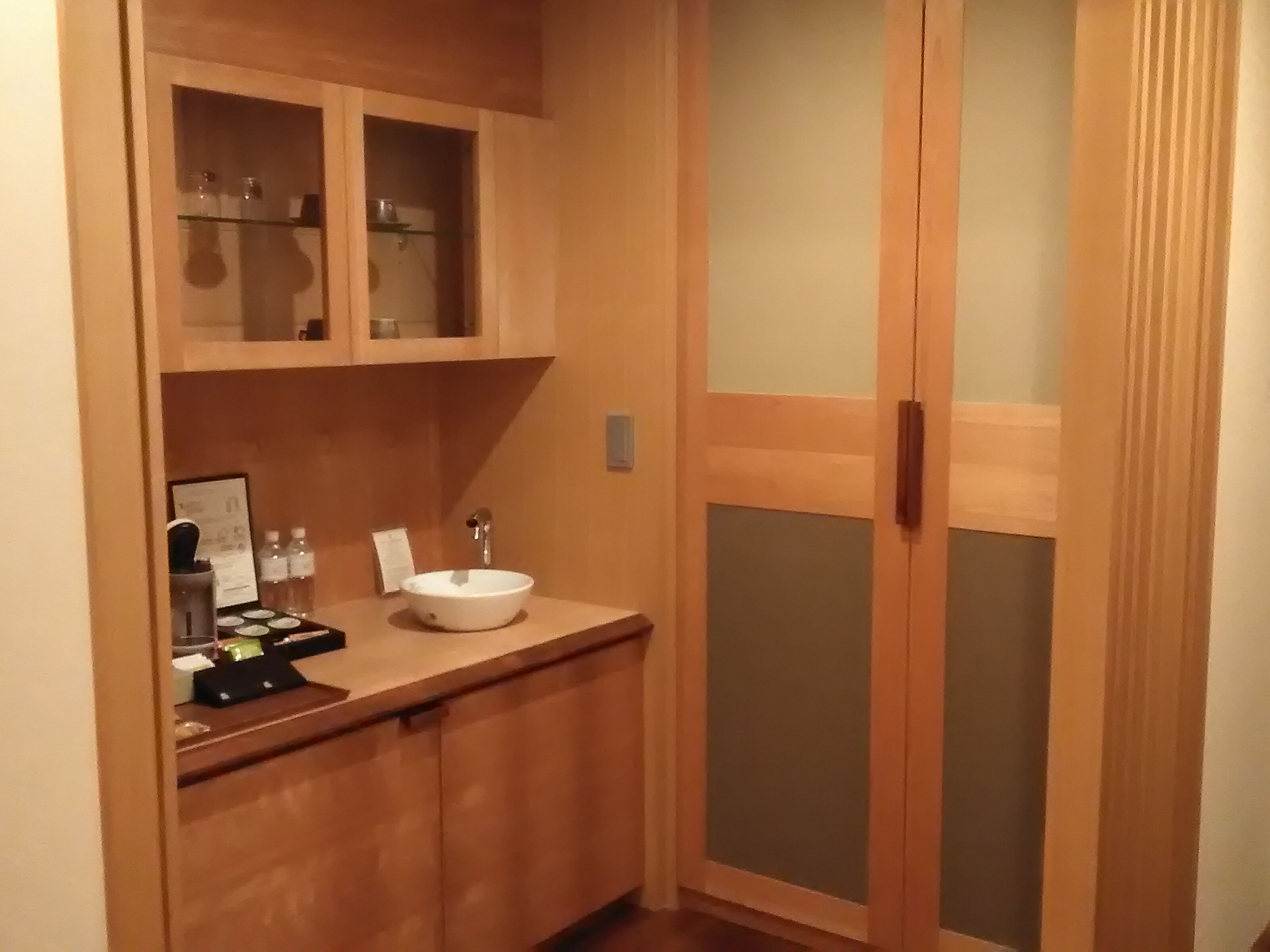 A small bathroom with a wooden vanity, a sink, and a mirror. There are cabinets above the vanity and a frosted glass door to the right.