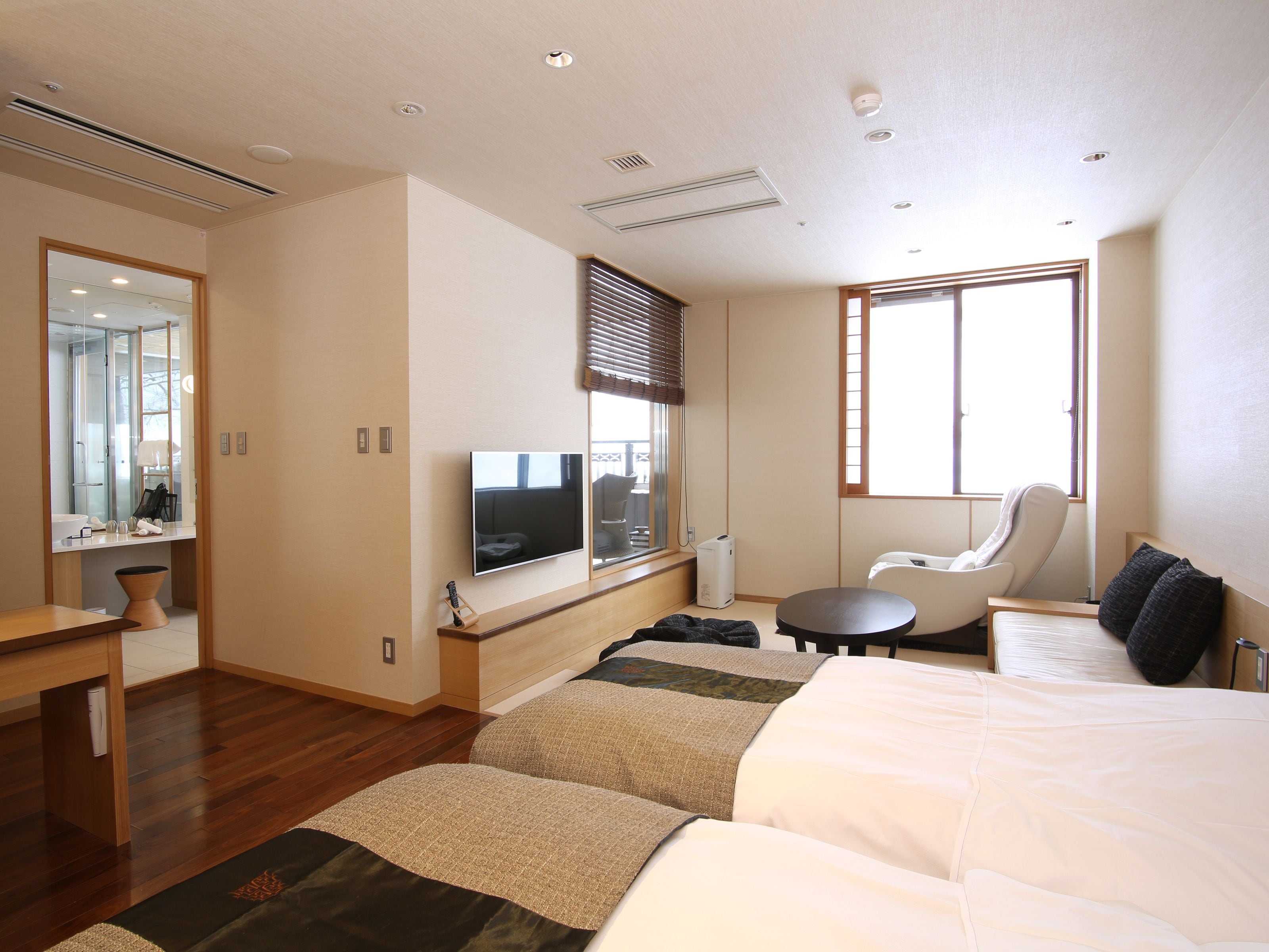 A modern hotel room with a large bed, a seating area, a flat-screen TV, and a window with blinds.