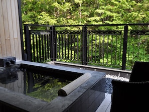 A hot tub with a wooden bench and a stone wall, surrounded by a black metal fence and lush greenery.