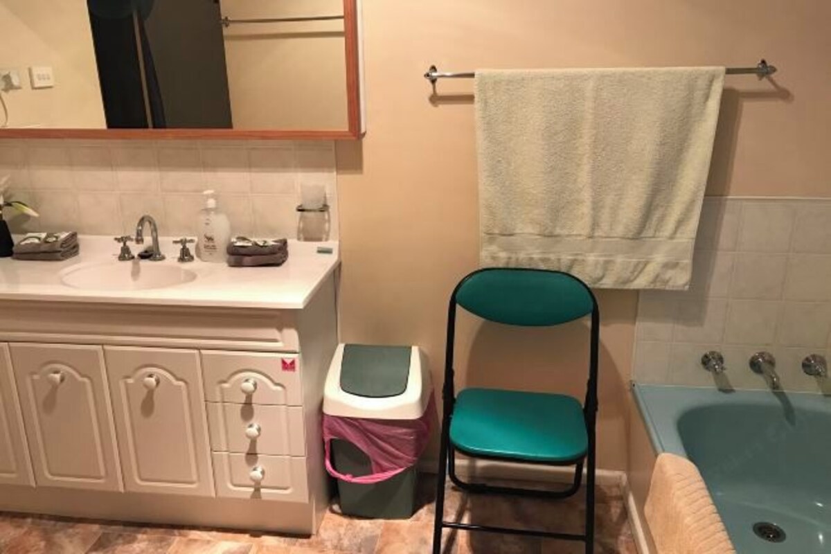 A bathroom with a sink, mirror, towel rack, chair, trash bin, and bathtub.
