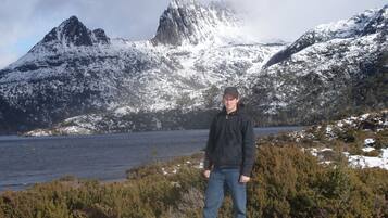 A person standing in a natural landscape with snow-covered mountains and a body of water in the background.