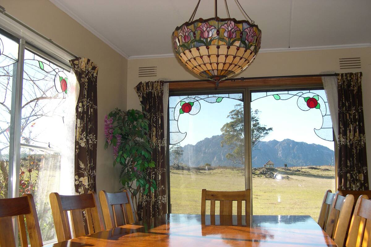 Un comedor con una gran mesa de madera, sillas, una lámpara de araña de vitrales colgante y vistas a un paisaje montañoso a través de amplios ventanales.
