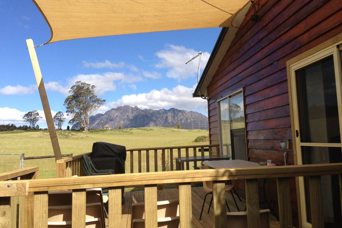Una terraza de madera con una mesa y sillas, una parrilla y vistas a montañas al fondo.