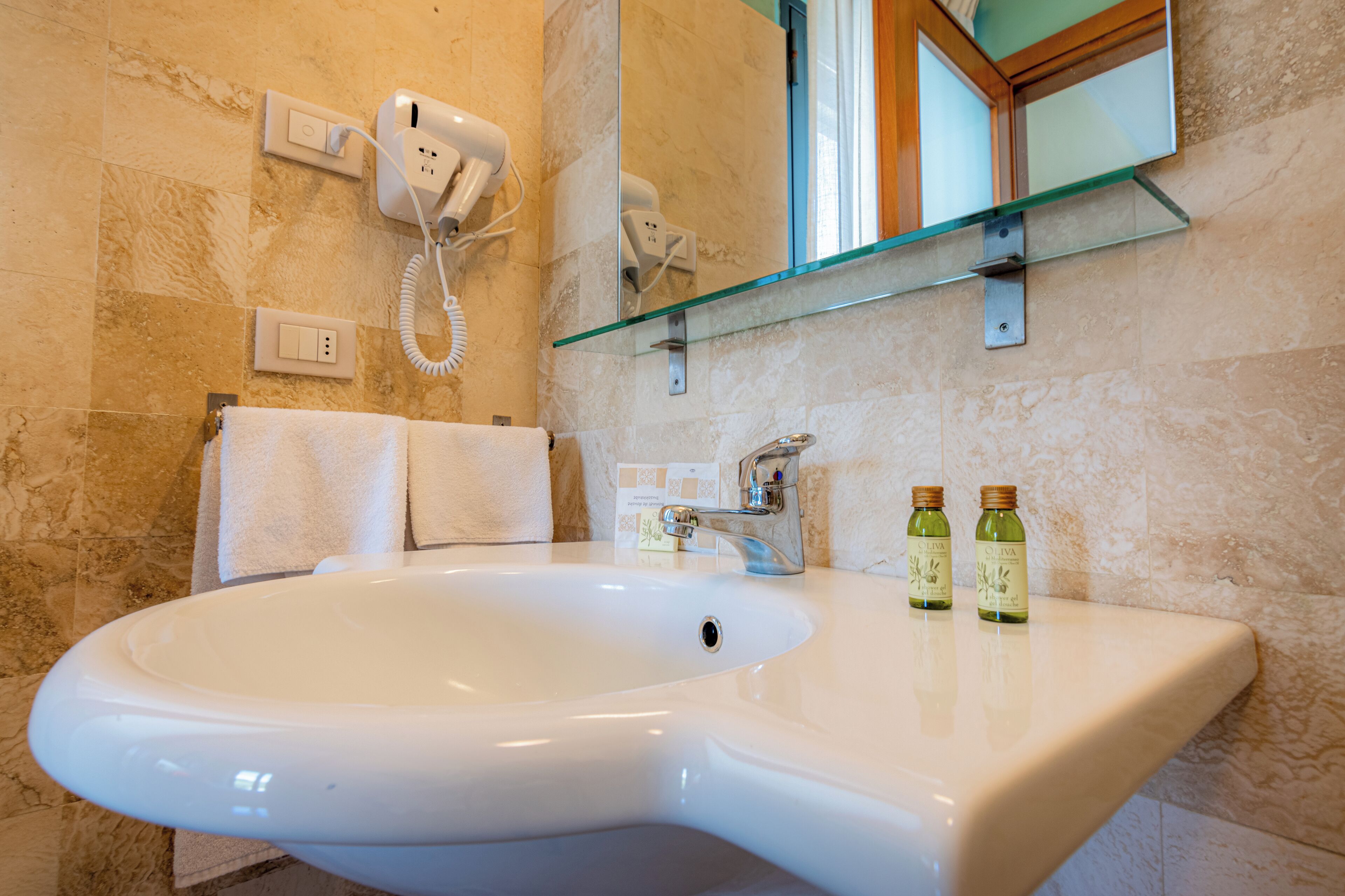 A bathroom with a large white sink, a glass shelf, a hairdryer, and two bottles of olive oil.