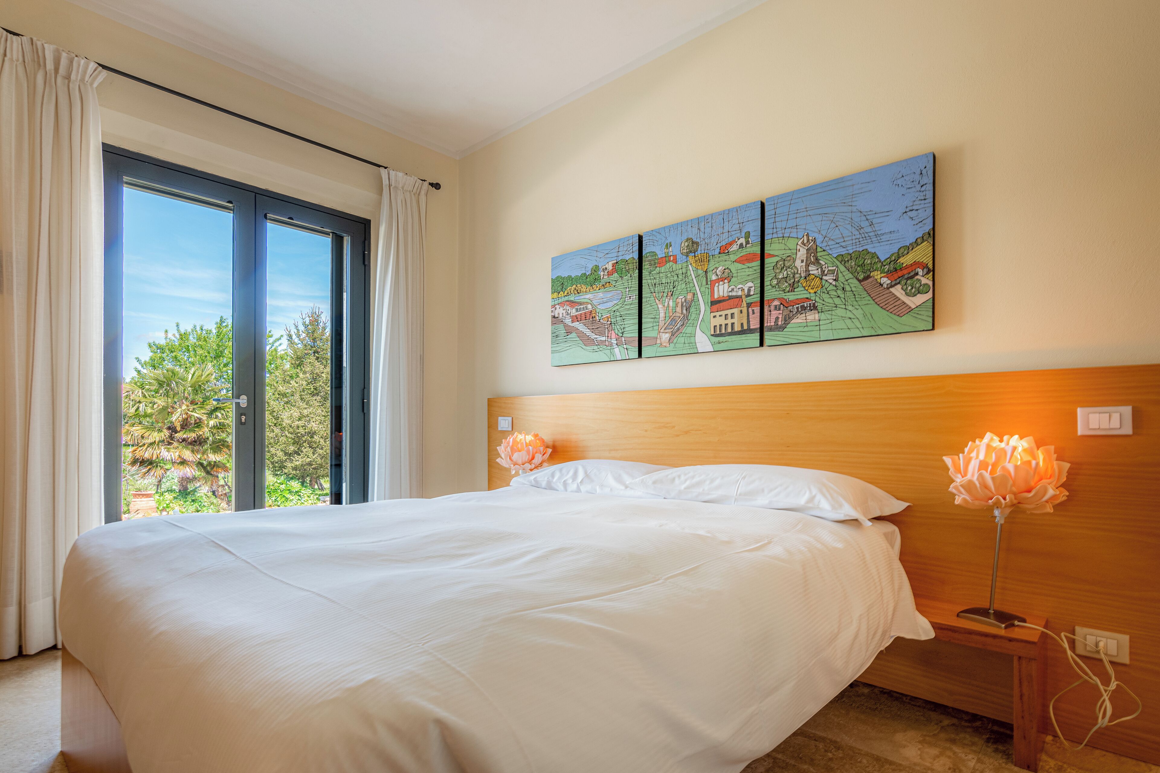A hotel room with a large bed, a painting on the wall, and a view through the window.