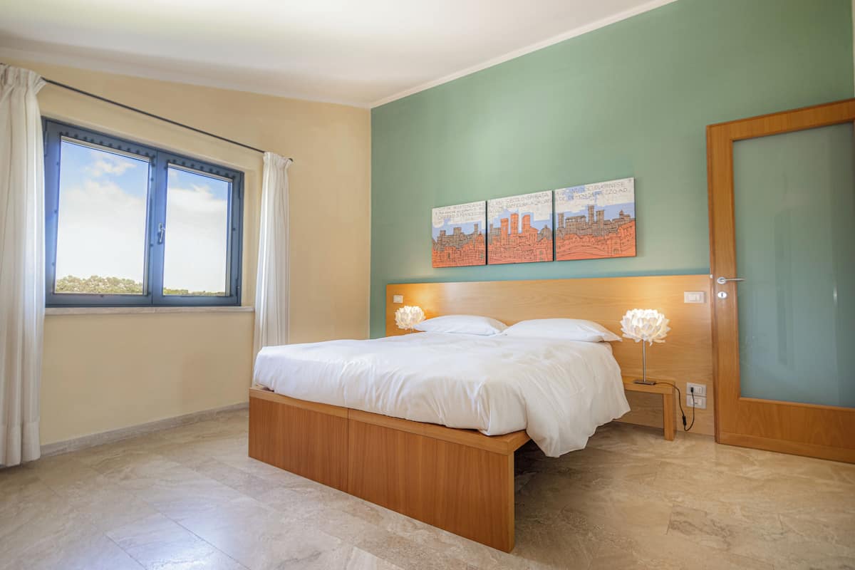 A bedroom with a bed, a window with curtains, a nightstand, and wall art.