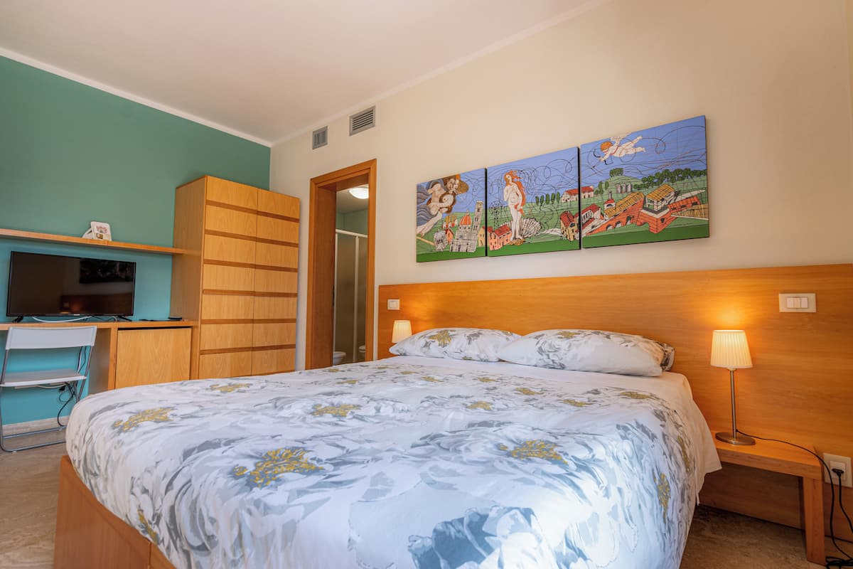A bedroom with a bed, a desk, a television, and a painting on the wall.