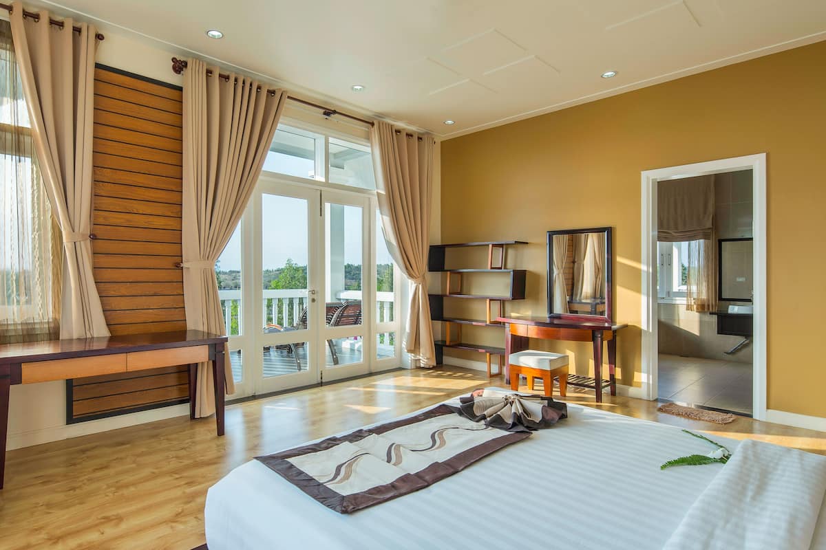 Ein Hotelzimmer mit einem großen Bett, einem Holzkopfteil, einem Schreibtisch, einem Sessel, einem kleinen Tisch, einem Spiegel und Blick ins Freie.