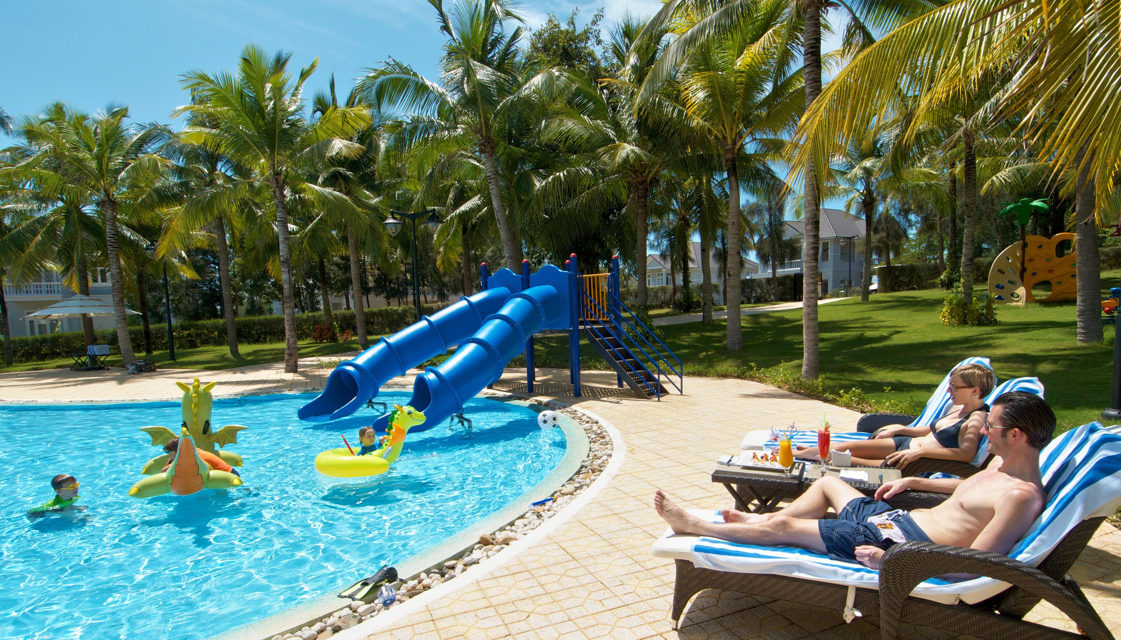 Una piscina con tobogán, sillas de descanso y palmeras.
