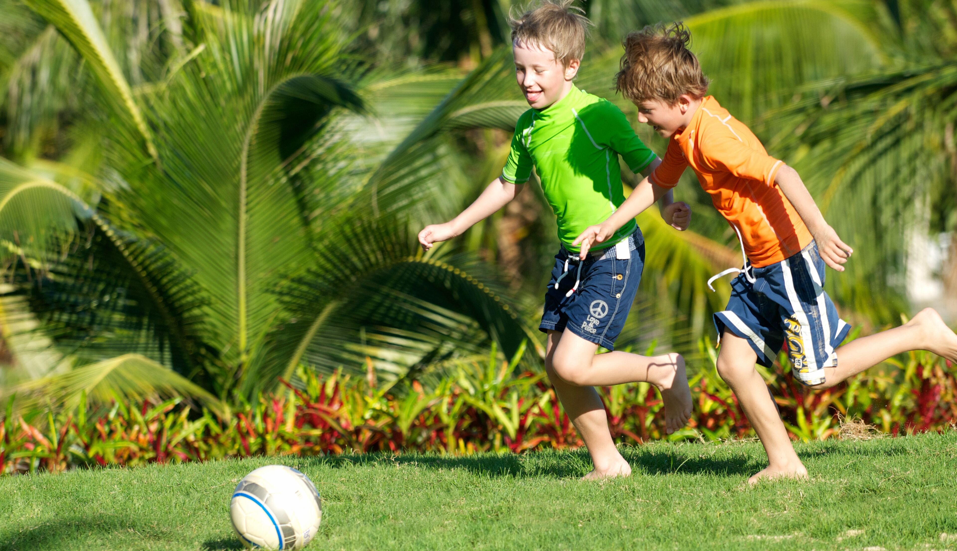Two children playing soccer on a grassy field with palm trees and colorful plants in the background.