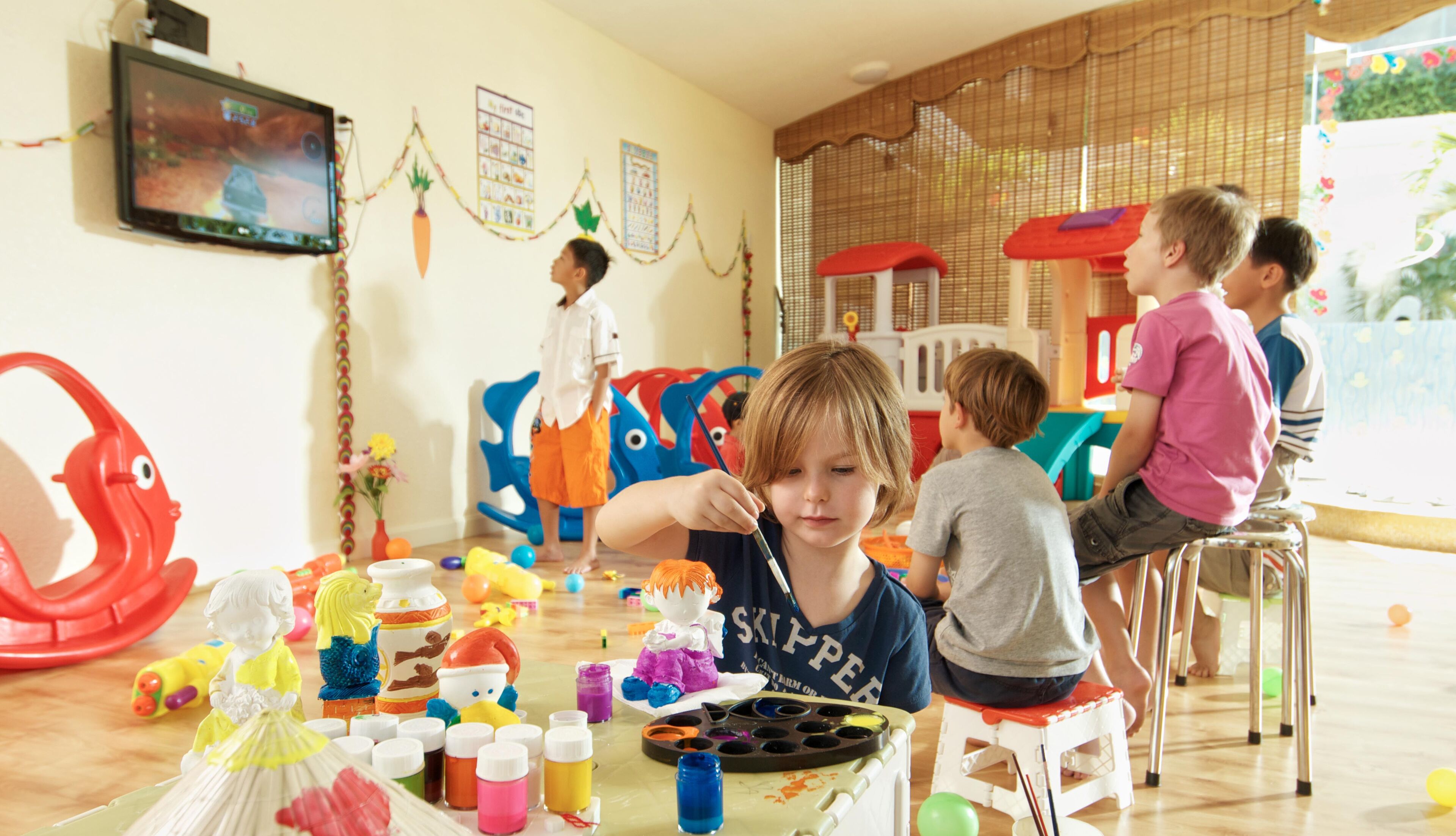 A room with a television mounted on the wall, a play area with colorful toys, and a table with art supplies.