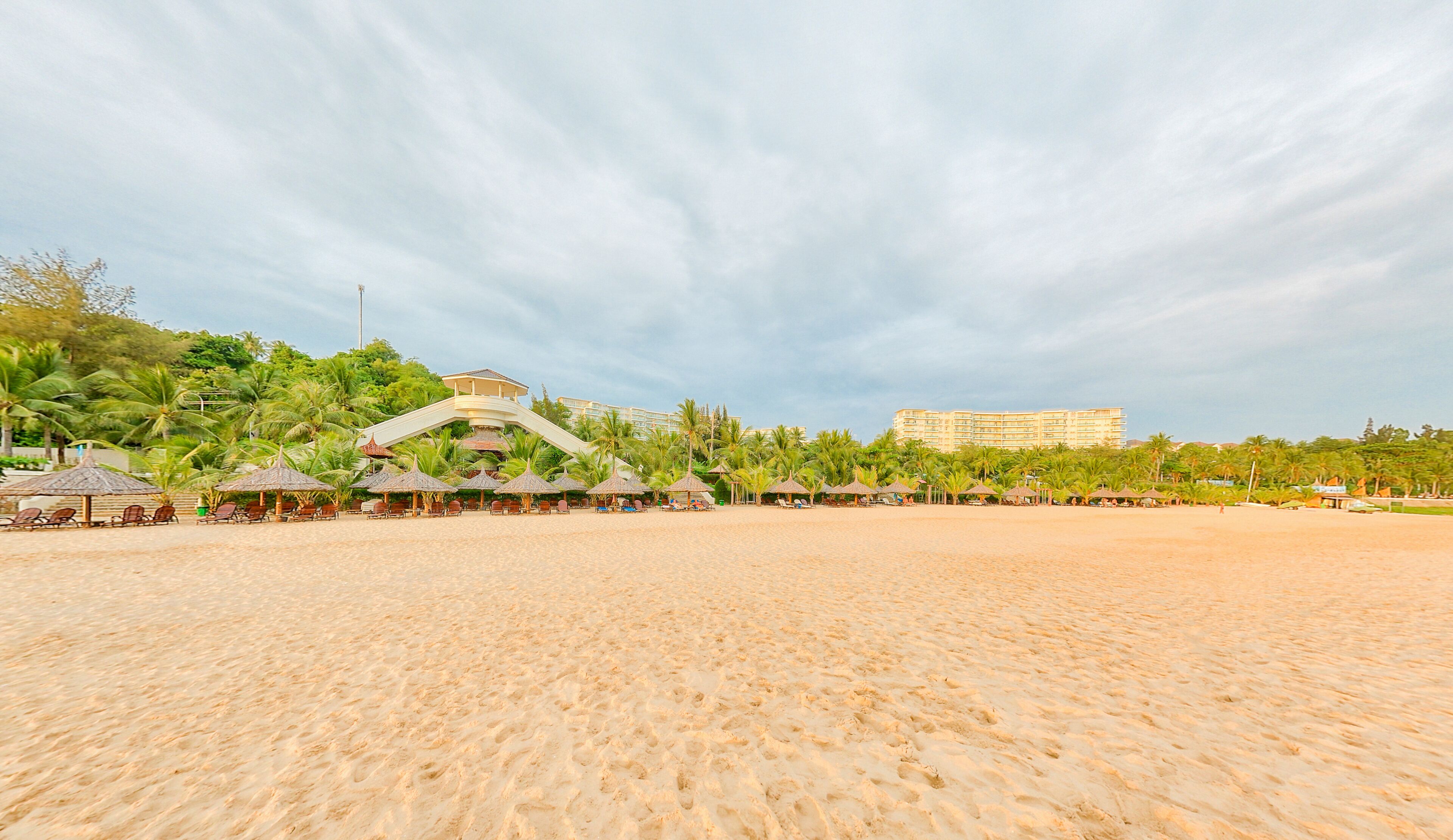 A beach with a large resort building in the background, palm trees, and beach umbrellas.