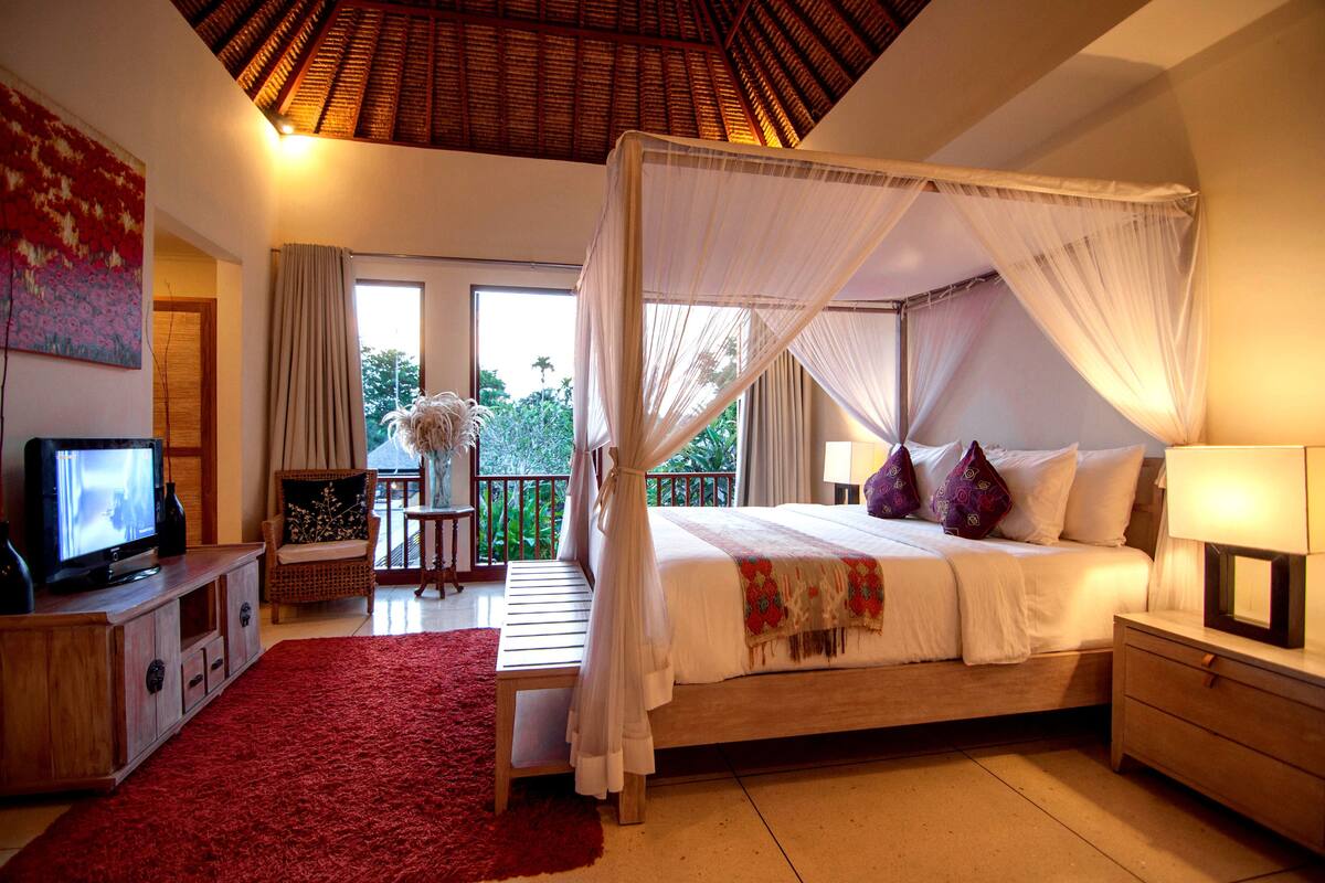 A bedroom with a four-poster bed, a television on a wooden cabinet, and a red rug.