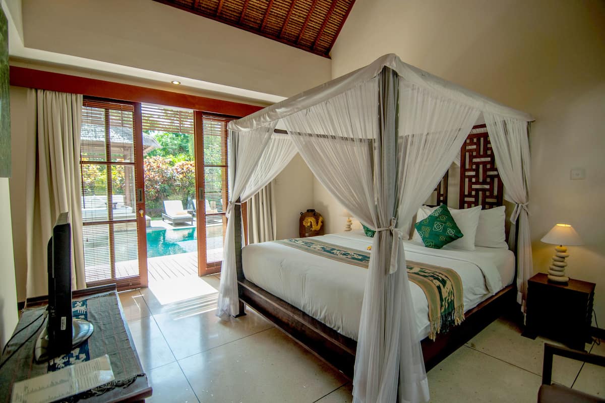 A room with a four-poster bed, a television on a stand, and a view of a pool and greenery outside.