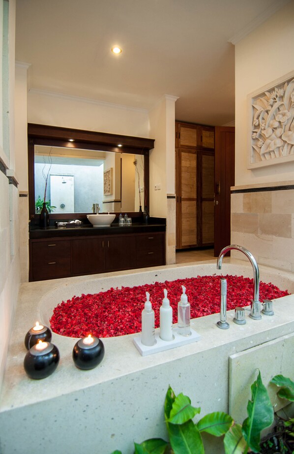 Une salle de bain avec une grande baignoire remplie de pétales de roses rouges, un meuble-lavabo en bois foncé et un grand miroir au-dessus du lavabo.